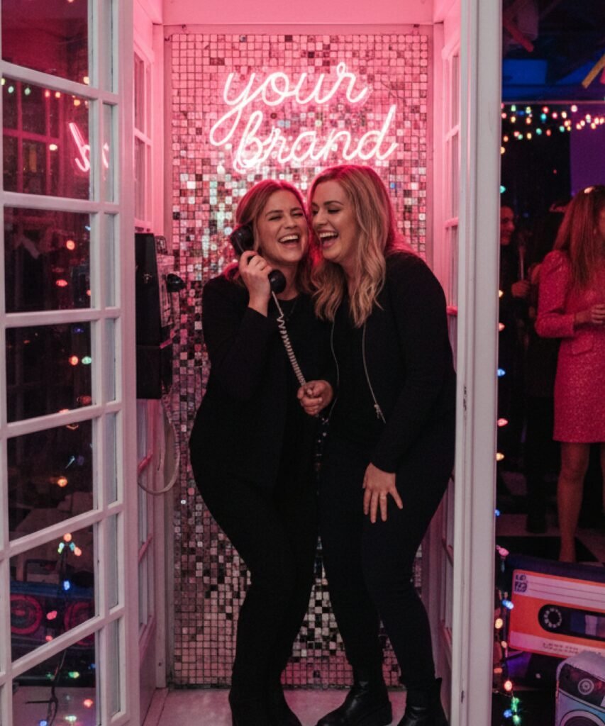 Cabine téléphonique blanche en soirée avec néon rose et fond sequins argentés.