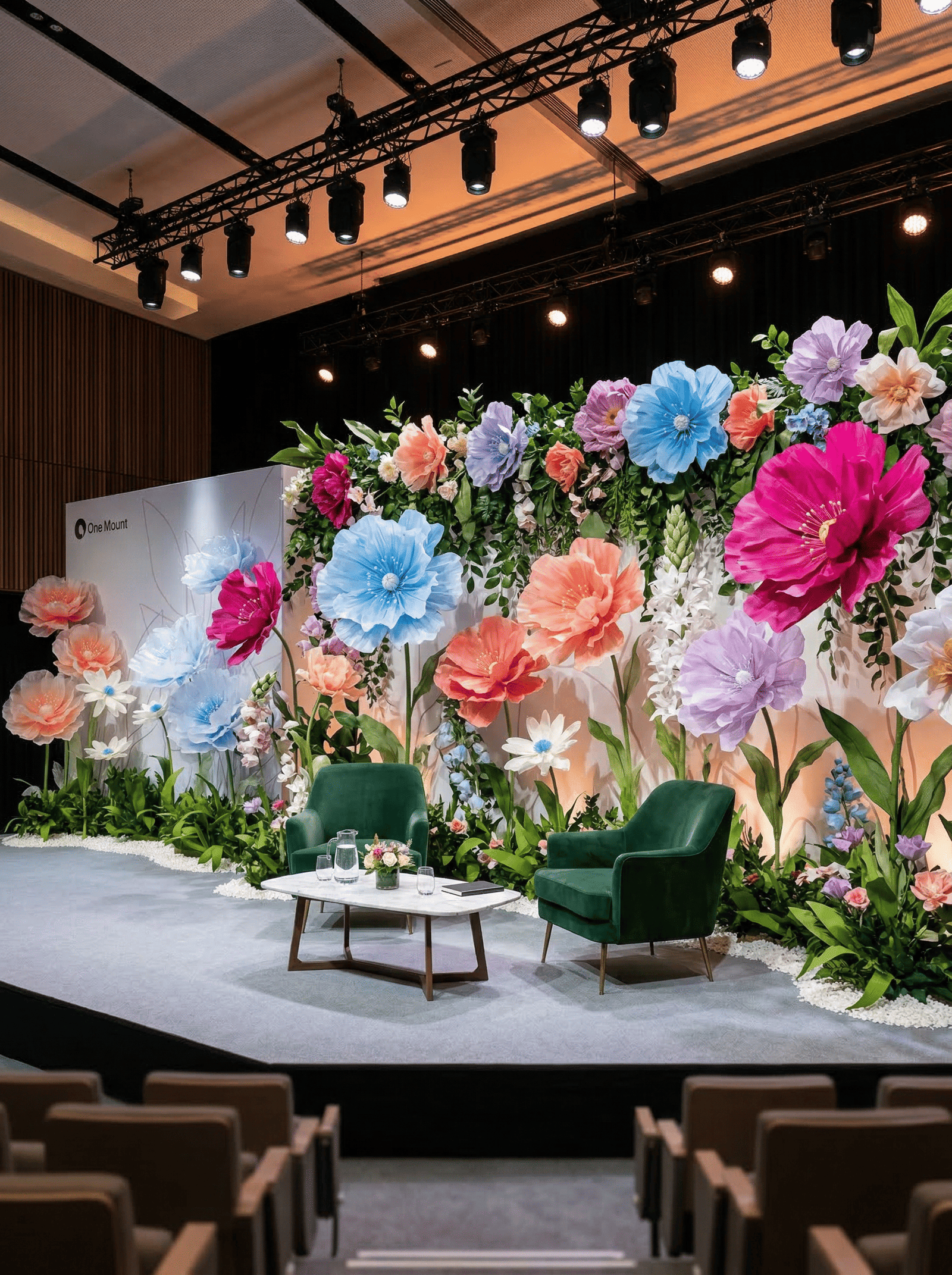Scène de conférence décorée avec fleurs géantes colorées et fauteuils pour table ronde.